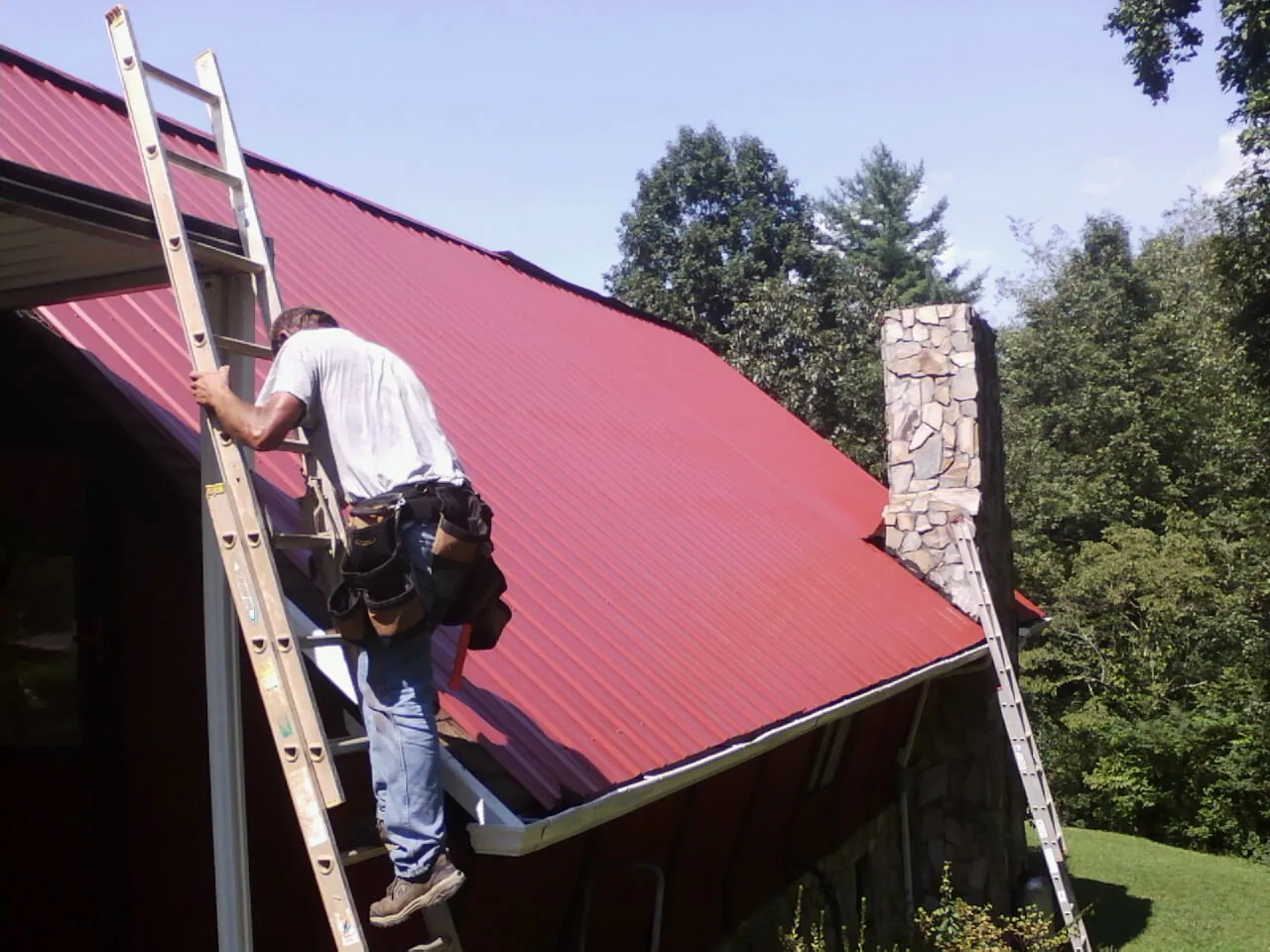 Roofer on a ladder beside a red metal roof and stone chimney — Soffit & Fascia Repair by Roofing Brothers in Dubois